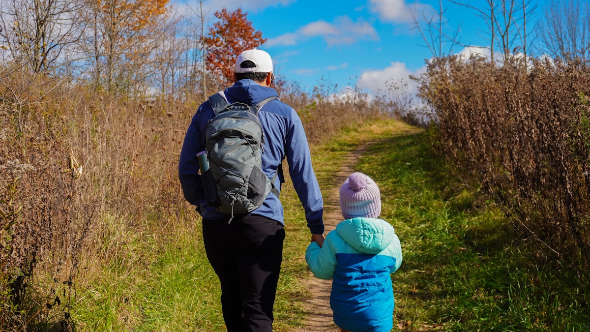 Parent carrying child in a backpack carrier on a forested trail, both looking at the surroundings