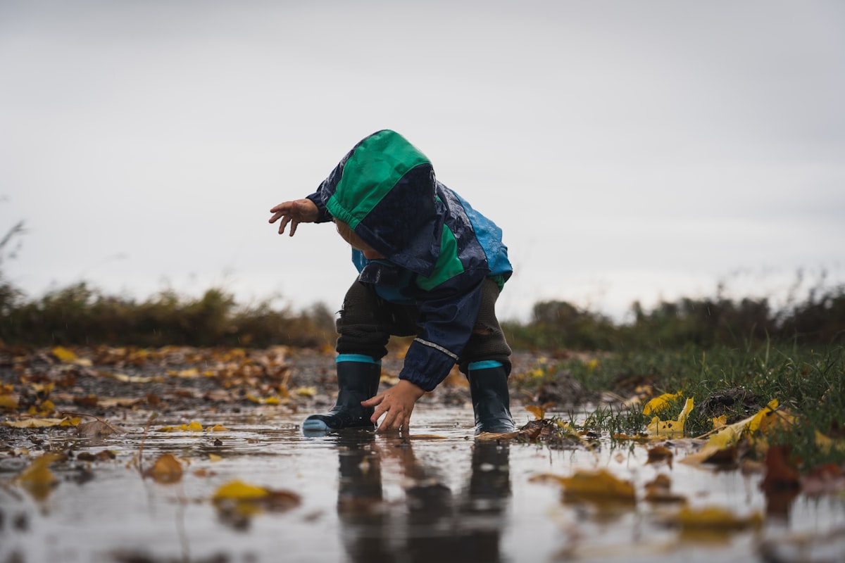 A 3 year old toddler exploring outdoors, crouching near a puddle in a backyard garden
