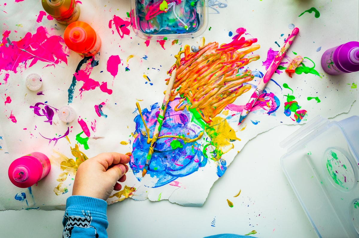 A preschool-age child engaged in open-ended painting and art play at a table, hands covered in colorful paint