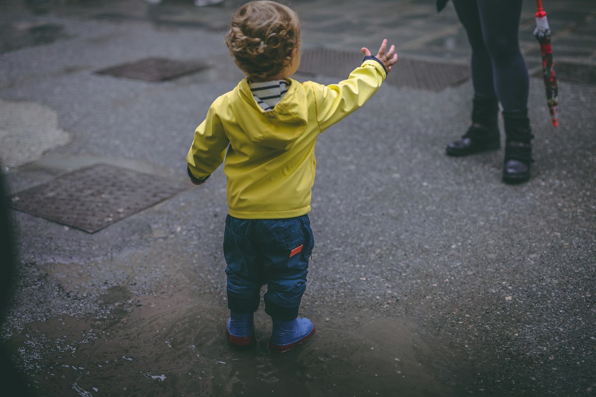 A toddler in a bright rain jacket stomping through a puddle on a wet trail