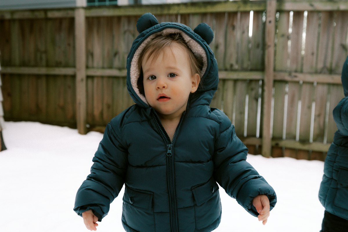 A toddler in a red snowsuit crouching down to touch fresh snow in a quiet winter yard