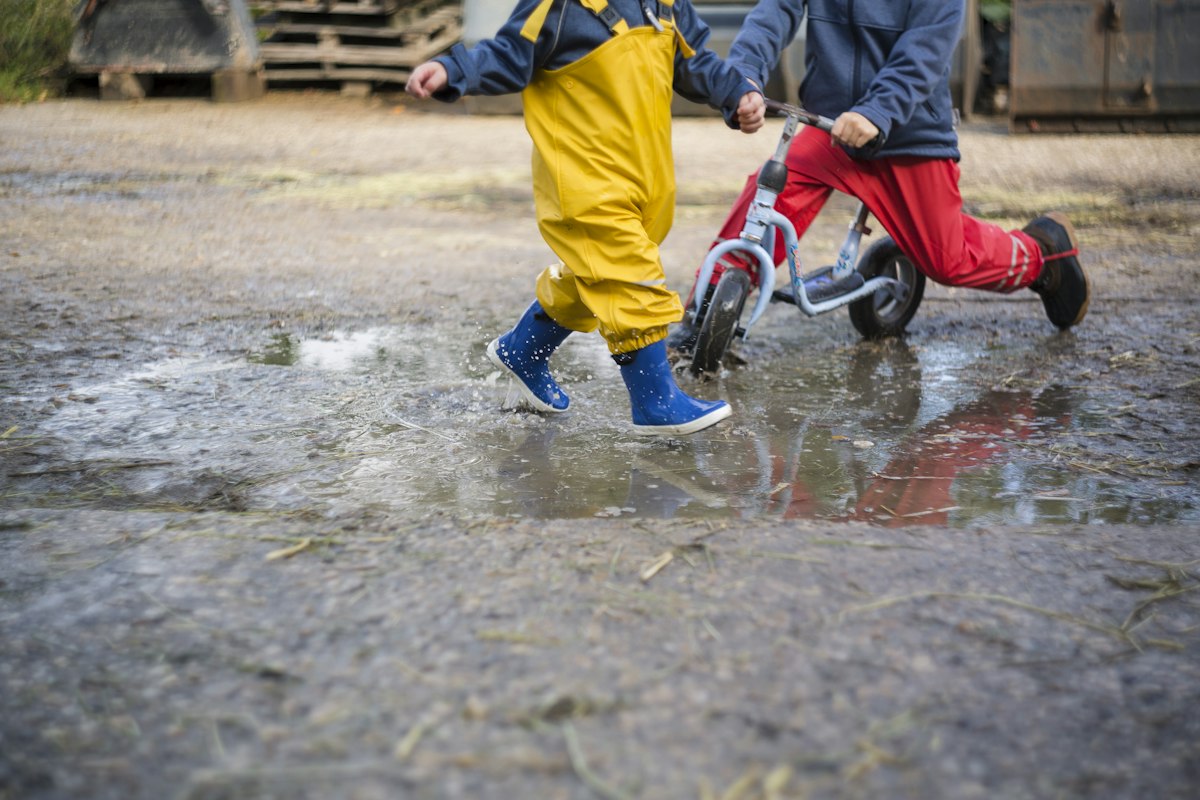 Two children in colourful rain boots jumping in a large puddle on a spring day