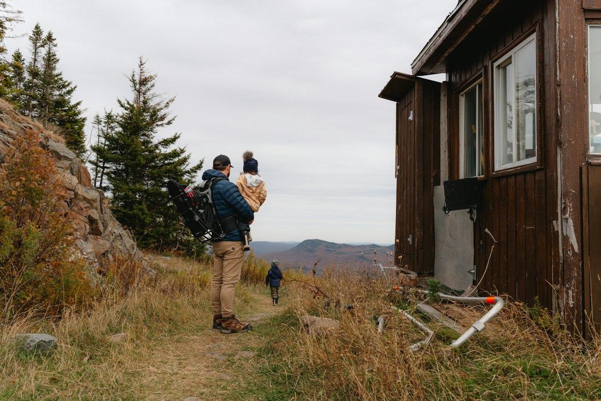 A parent hiking a sunny mountain trail with a young child in a framed backpack carrier